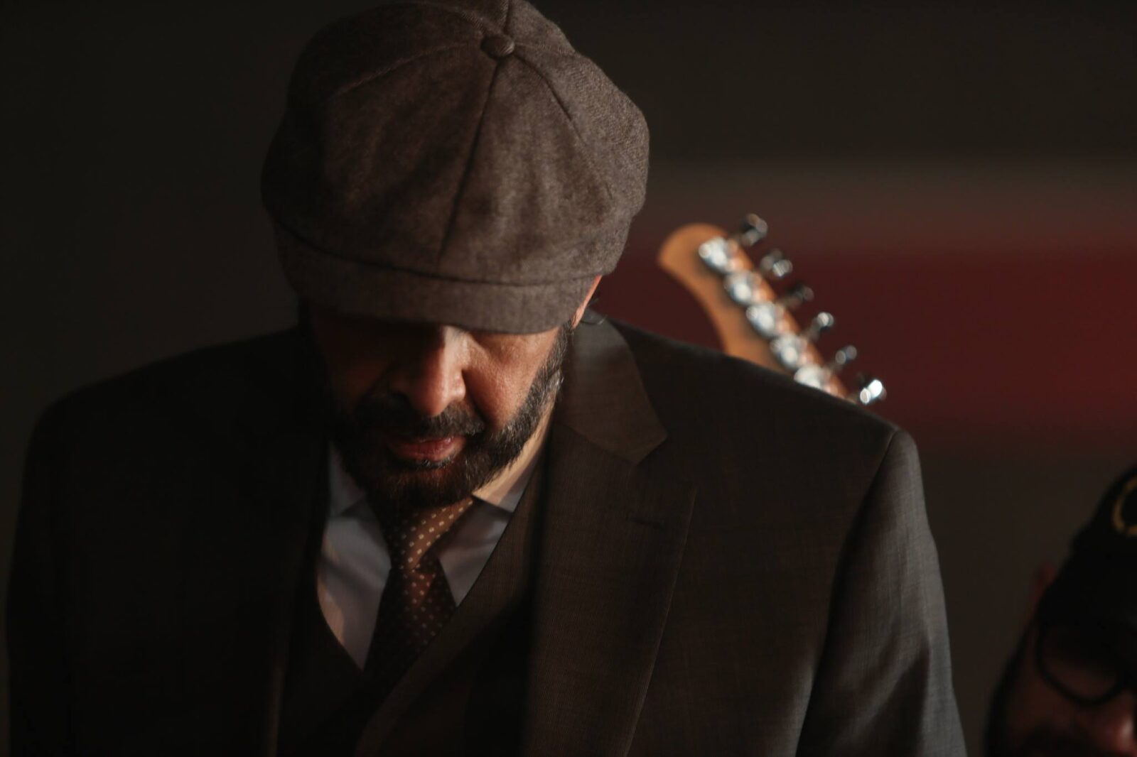 Man in a flat cap and suit in a dimly lit room with a guitar.