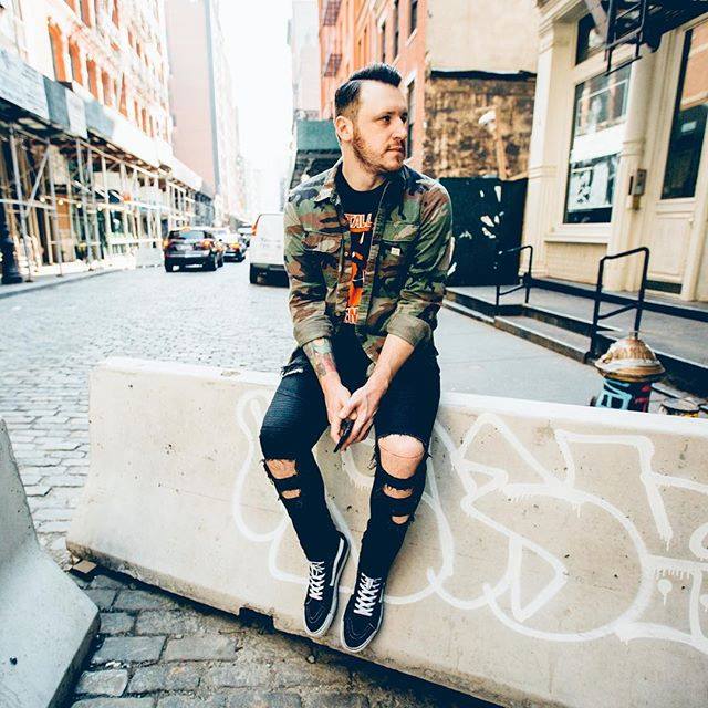 Young man with tattoos sitting on a graffiti-covered concrete block in an urban setting.