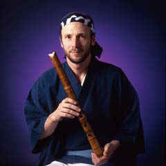 Man in traditional attire holding a wooden flute against a dark background.