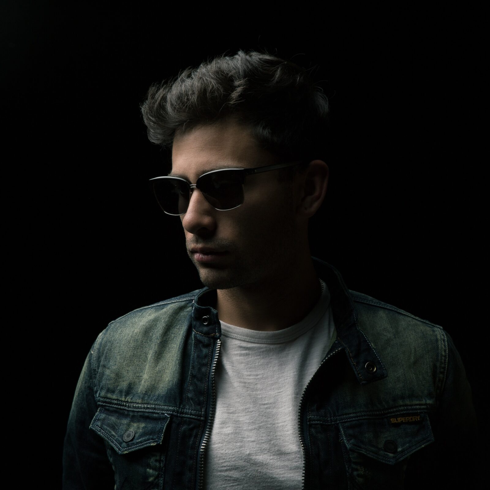 Man wearing sunglasses and a denim jacket in low light.