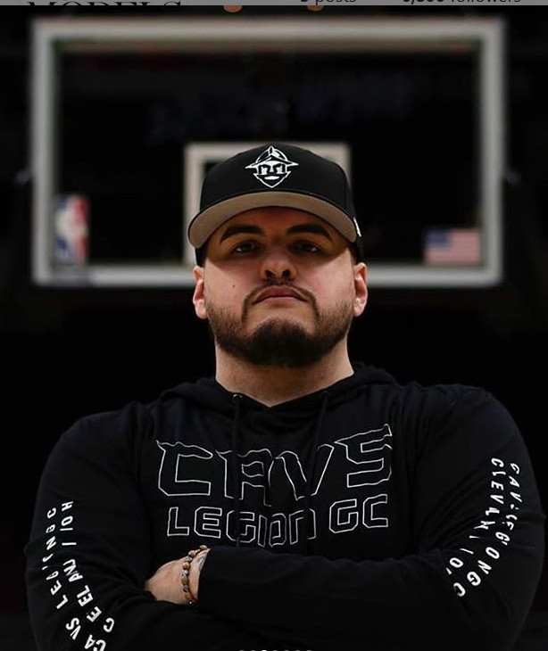 Confident man wearing a black hoodie and cap, standing with arms crossed in a basketball court.